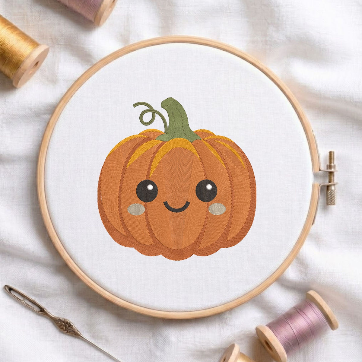 Cute smiling pumpkin machine embroidery design with a curly green stem and rosy cheeks, displayed inside a round wooden hoop on white linen fabric alongside thread spools and a needle.