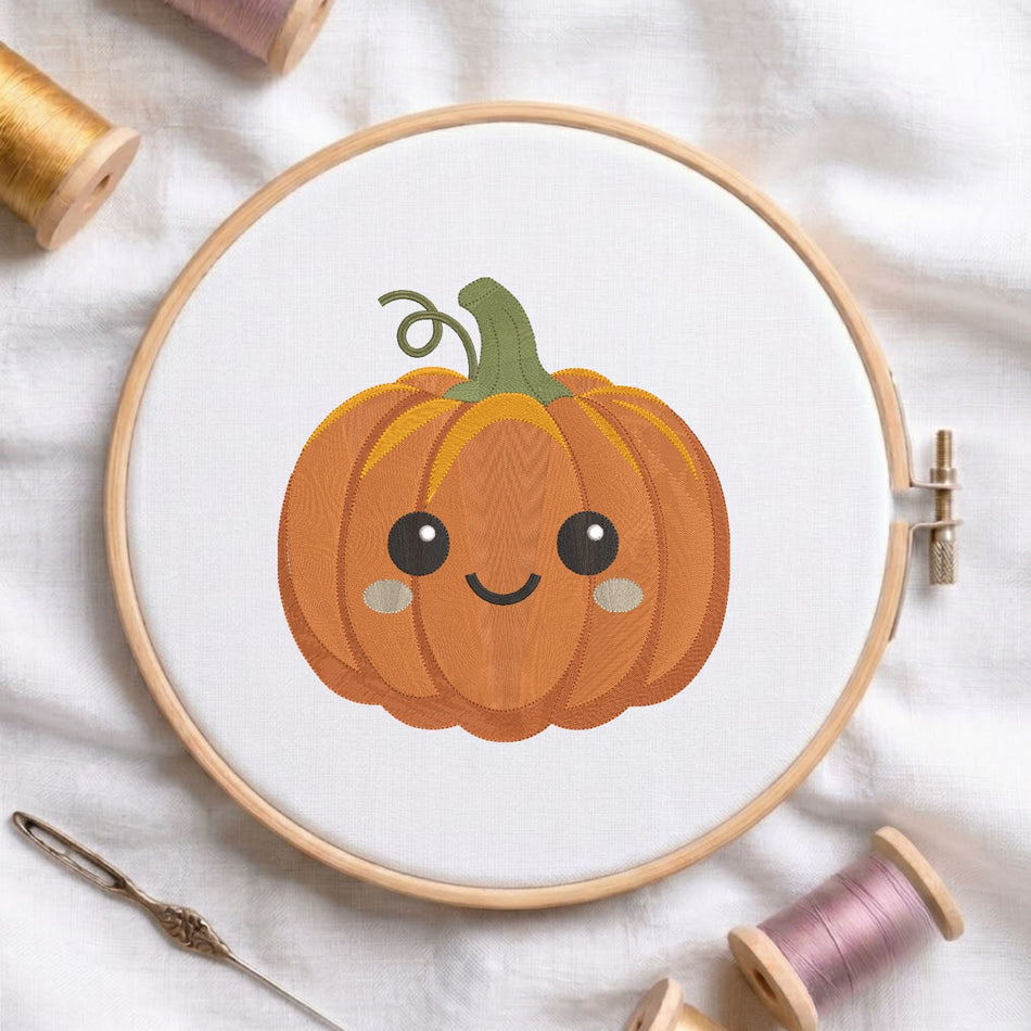 Cute smiling pumpkin machine embroidery design with a curly green stem and rosy cheeks, displayed inside a round wooden hoop on white linen fabric alongside thread spools and a needle.