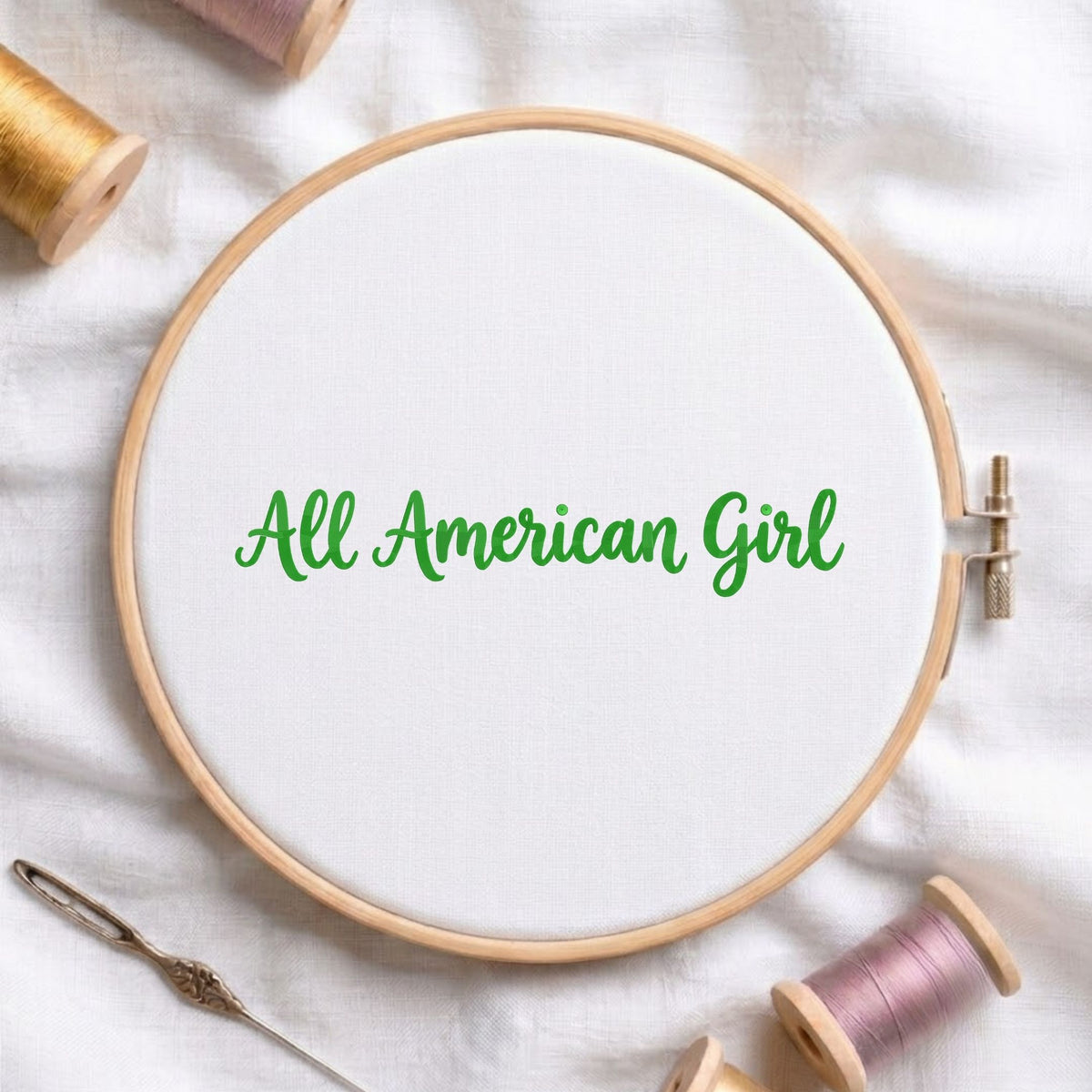 A wooden embroidery hoop holding white fabric with the phrase 'All American Girl' stitched in green script. The hoop is styled on a white fabric surface with spools of thread and a needle around it.