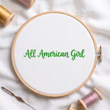 A wooden embroidery hoop holding white fabric with the phrase 'All American Girl' stitched in green script. The hoop is styled on a white fabric surface with spools of thread and a needle around it.