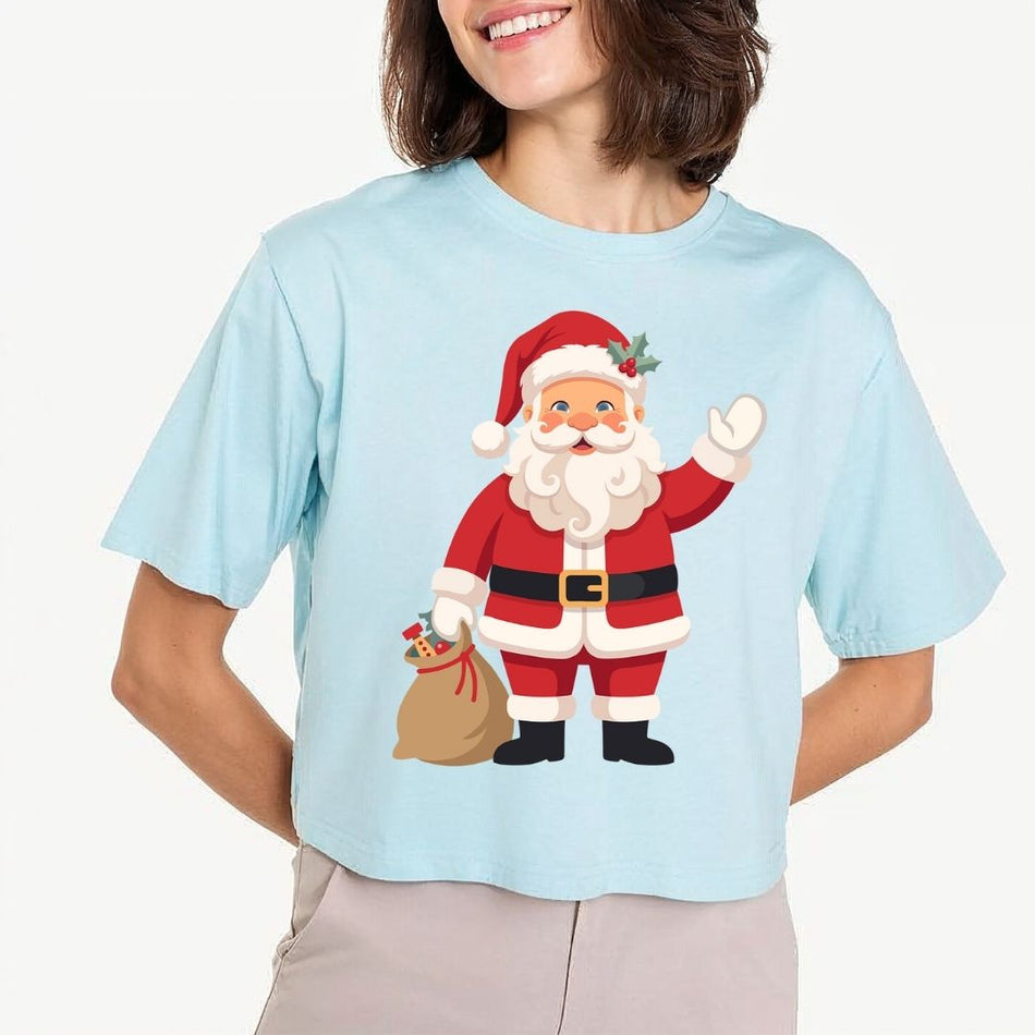 A woman wearing a light blue cropped t-shirt with a cartoon graphic of Santa Claus waving and holding a sack of gifts. The background is a clean, off-white studio setting.