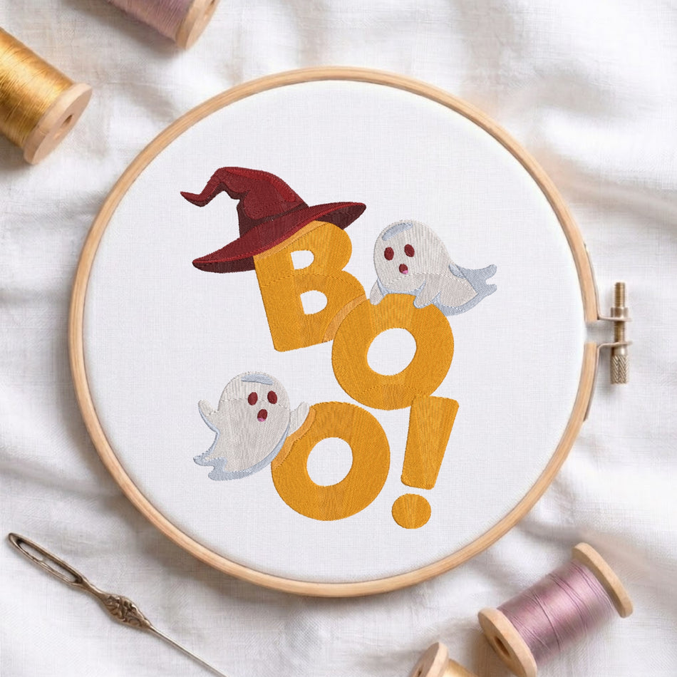 A white fabric stretched in a wooden embroidery hoop, featuring large embroidered orange "BOO!" letters, a burgundy witch hat perched on top, and two playful white ghosts.