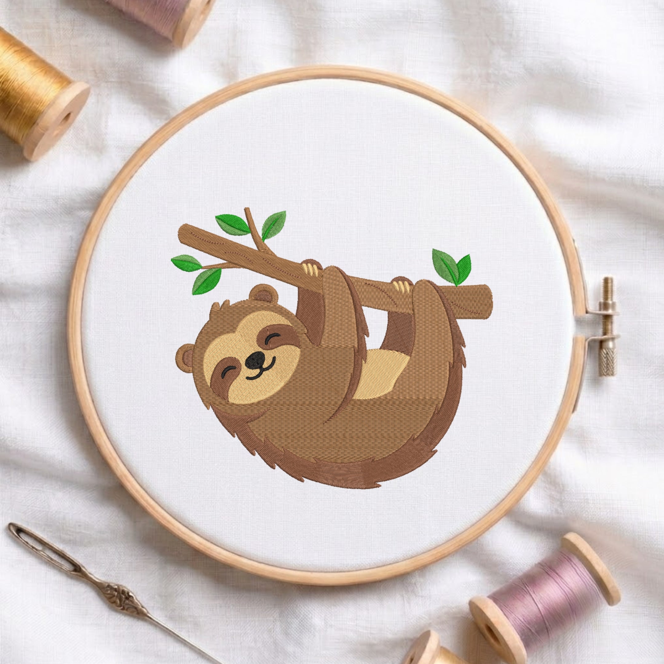 An embroidered design of a smiling brown sloth hanging upside down from a tree branch with green leaves, stitched inside a wooden hoop on white fabric with sewing threads.
