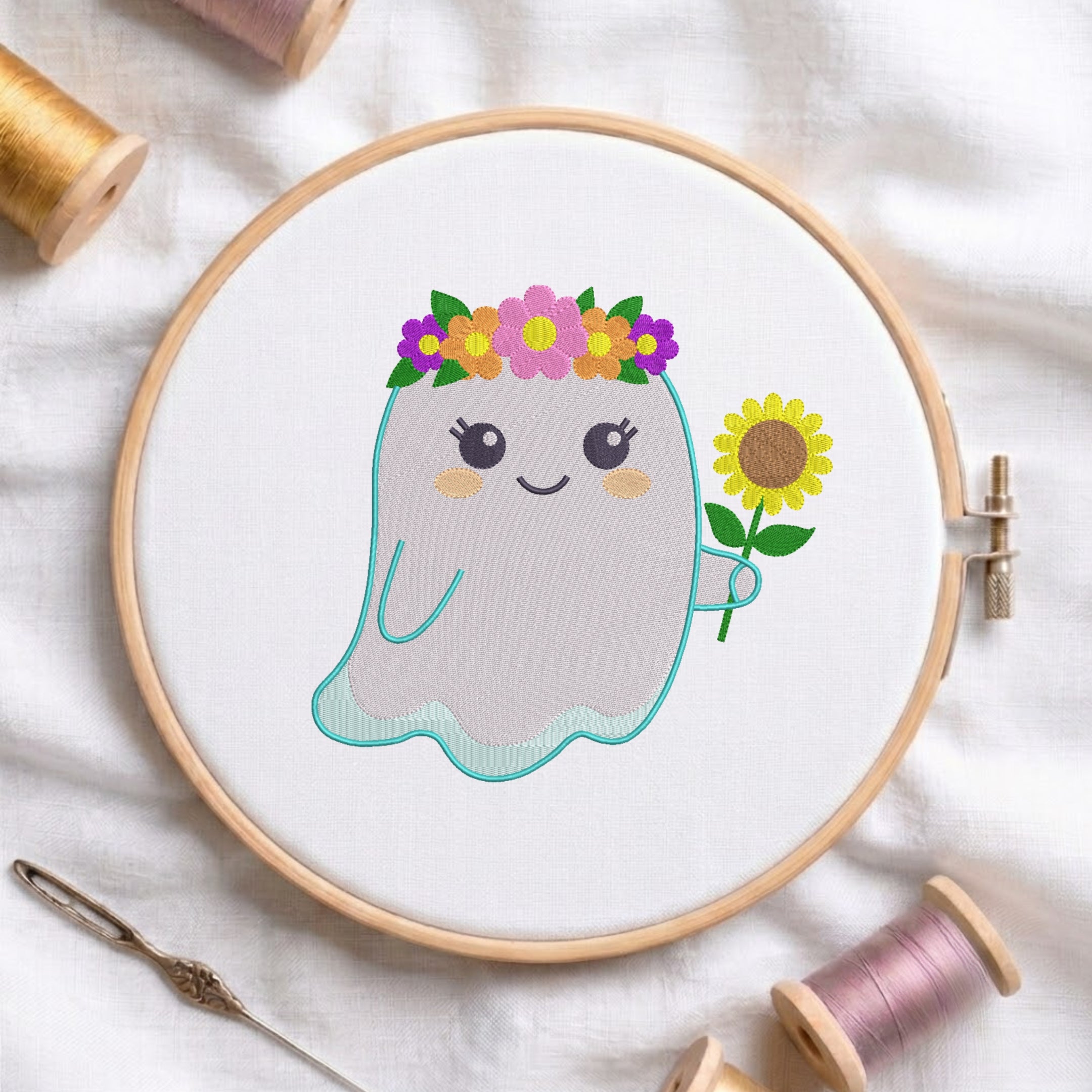 A circular embroidery hoop holding white fabric with a cute ghost design. The ghost has a teal stitched outline, a smiling face, a flower crown with pink, purple, yellow, and green flowers, and is holding a yellow sunflower.
