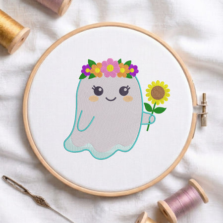 A circular embroidery hoop holding white fabric with a cute ghost design. The ghost has a teal stitched outline, a smiling face, a flower crown with pink, purple, yellow, and green flowers, and is holding a yellow sunflower.