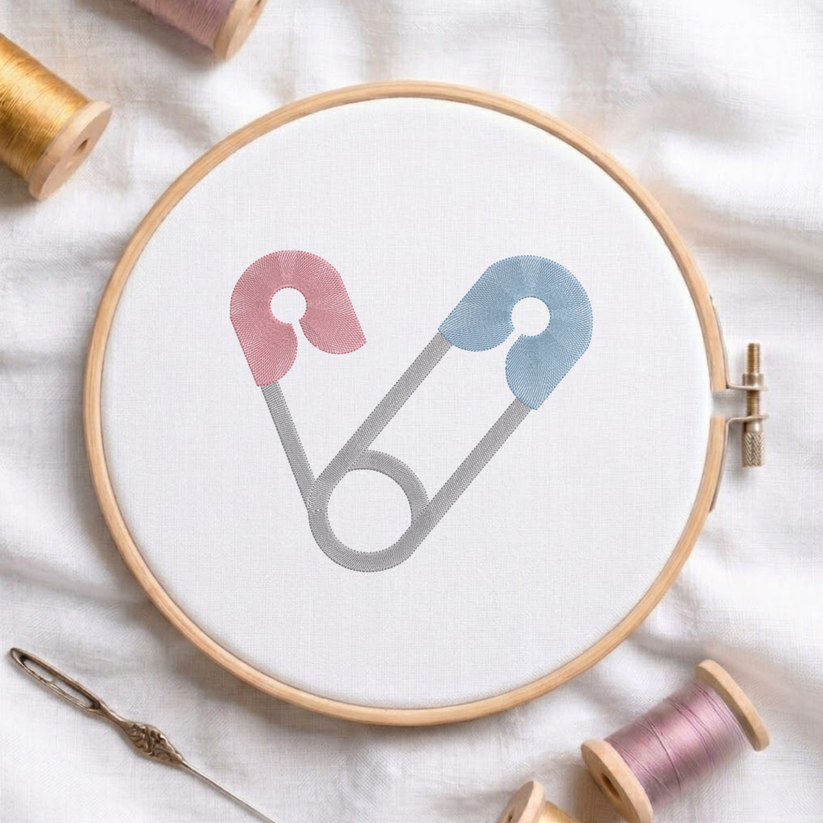 A circular embroidery hoop holding white fabric, with a minimalist safety-pin style design in pink, blue, and gray stitched in the center. The hoop is styled on a white fabric background with spools of thread and a needle around it.
