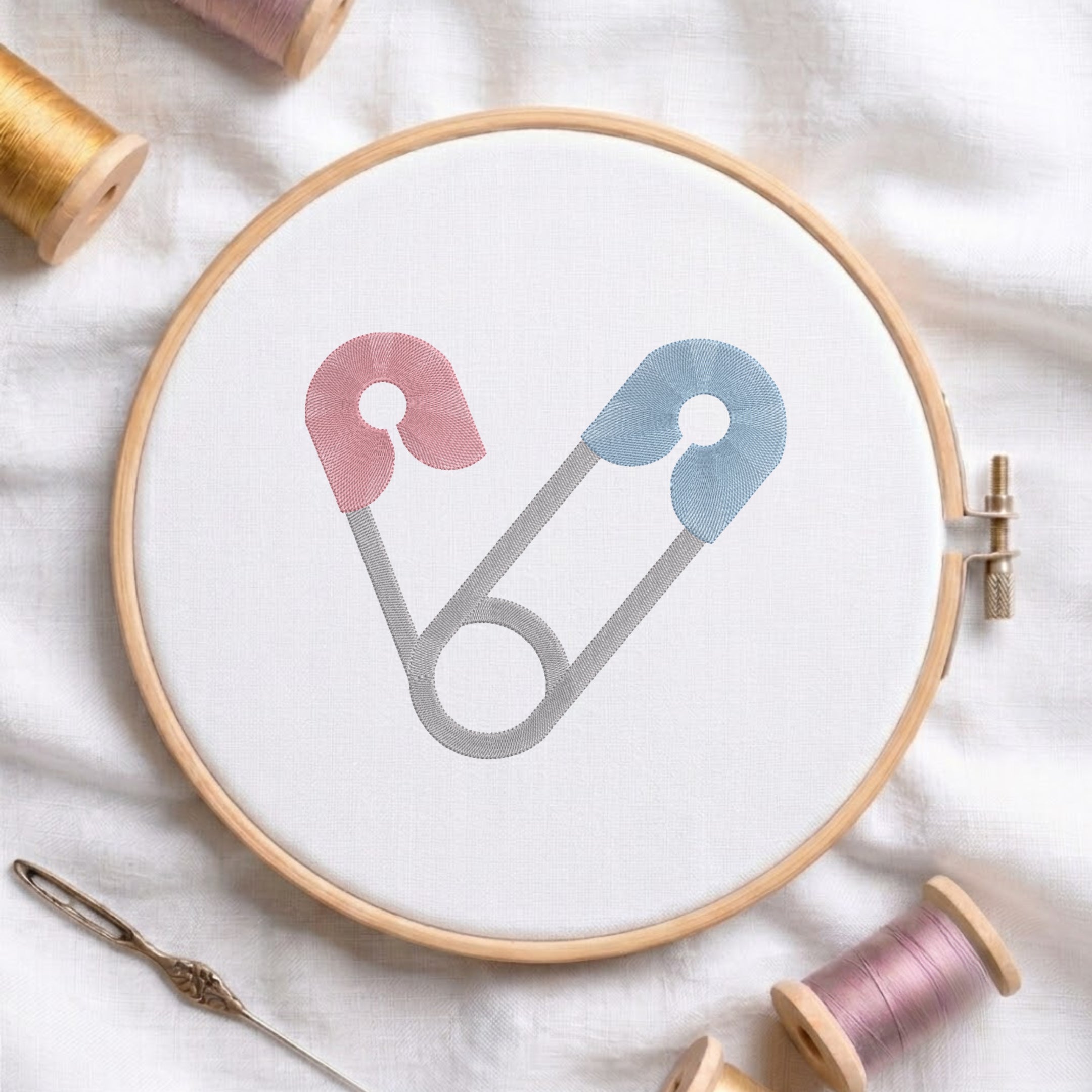 A circular embroidery hoop holding white fabric, with a minimalist safety-pin style design in pink, blue, and gray stitched in the center. The hoop is styled on a white fabric background with spools of thread and a needle around it.