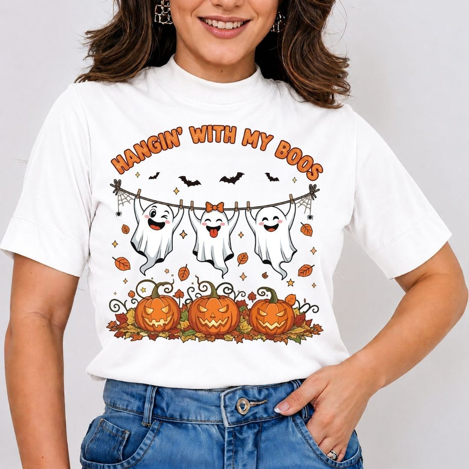 A woman wearing a white short-sleeve t-shirt featuring three friendly cartoon ghosts hanging from a clothesline above carved pumpkins with the text "HANGIN' WITH MY BOOS."