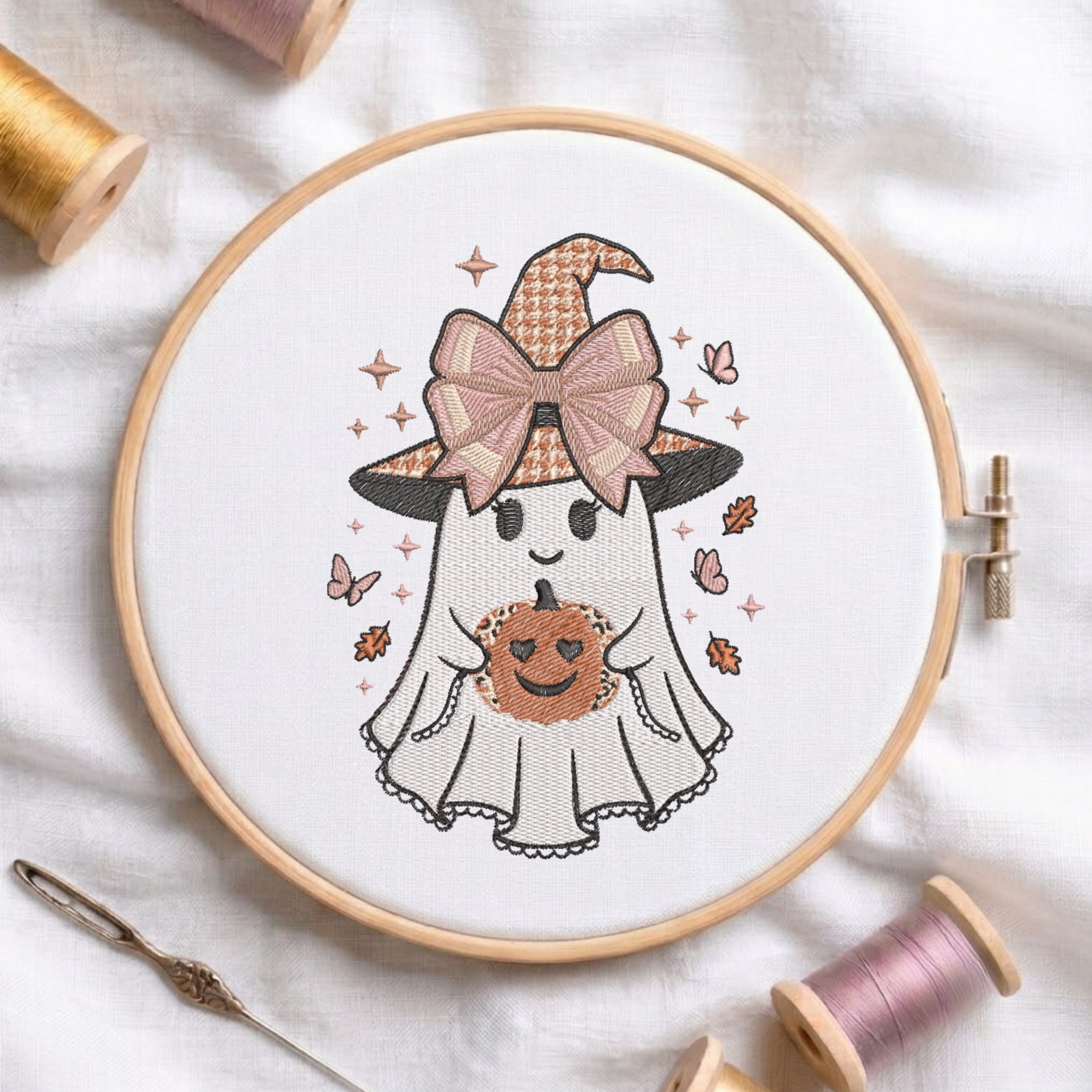 A circular embroidery hoop displays a cute cartoon ghost holding a pumpkin with a witch hat and bow. The hoop is styled on a white fabric background with spools of thread and a needle.