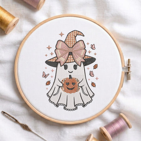 A circular embroidery hoop displays a cute cartoon ghost holding a pumpkin with a witch hat and bow. The hoop is styled on a white fabric background with spools of thread and a needle.