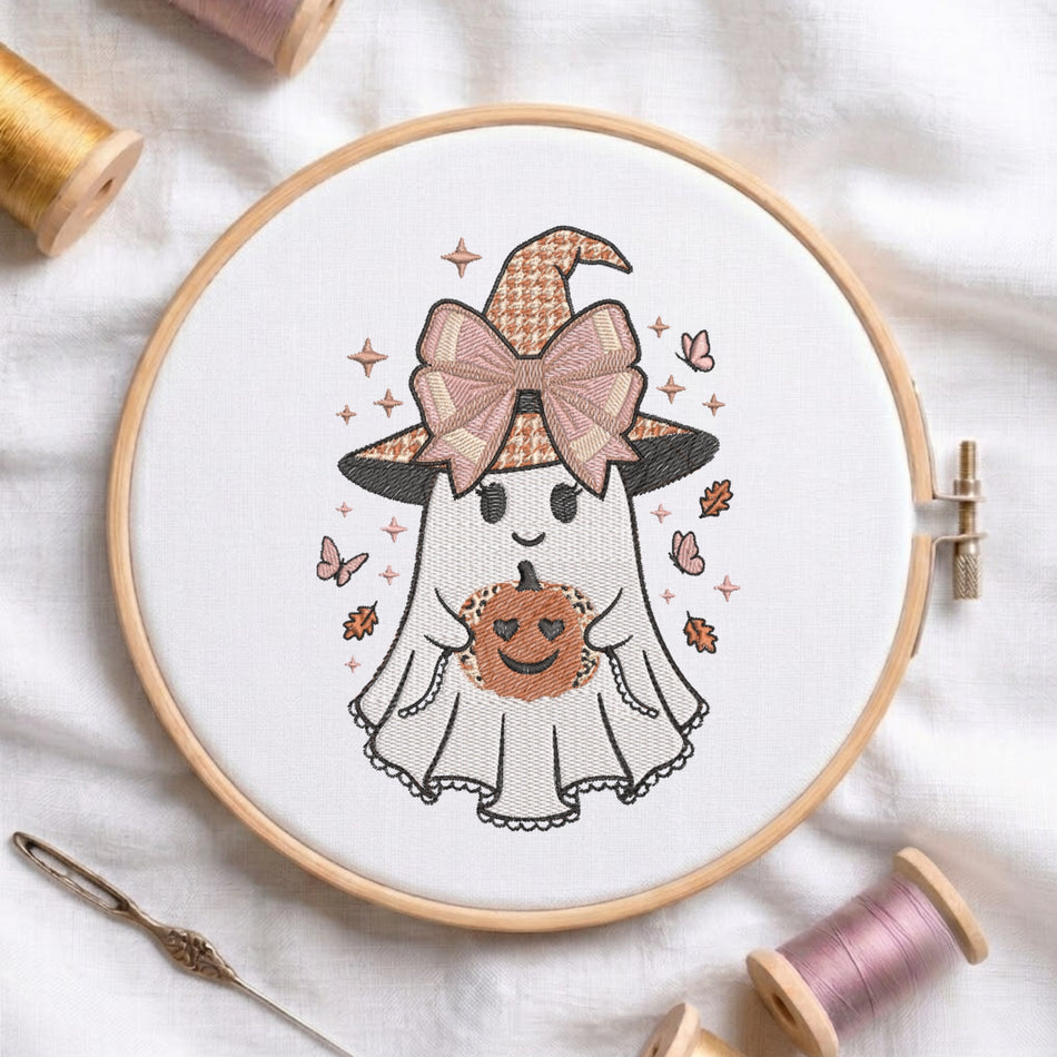 A circular embroidery hoop displays a cute cartoon ghost holding a pumpkin with a witch hat and bow. The hoop is styled on a white fabric background with spools of thread and a needle.