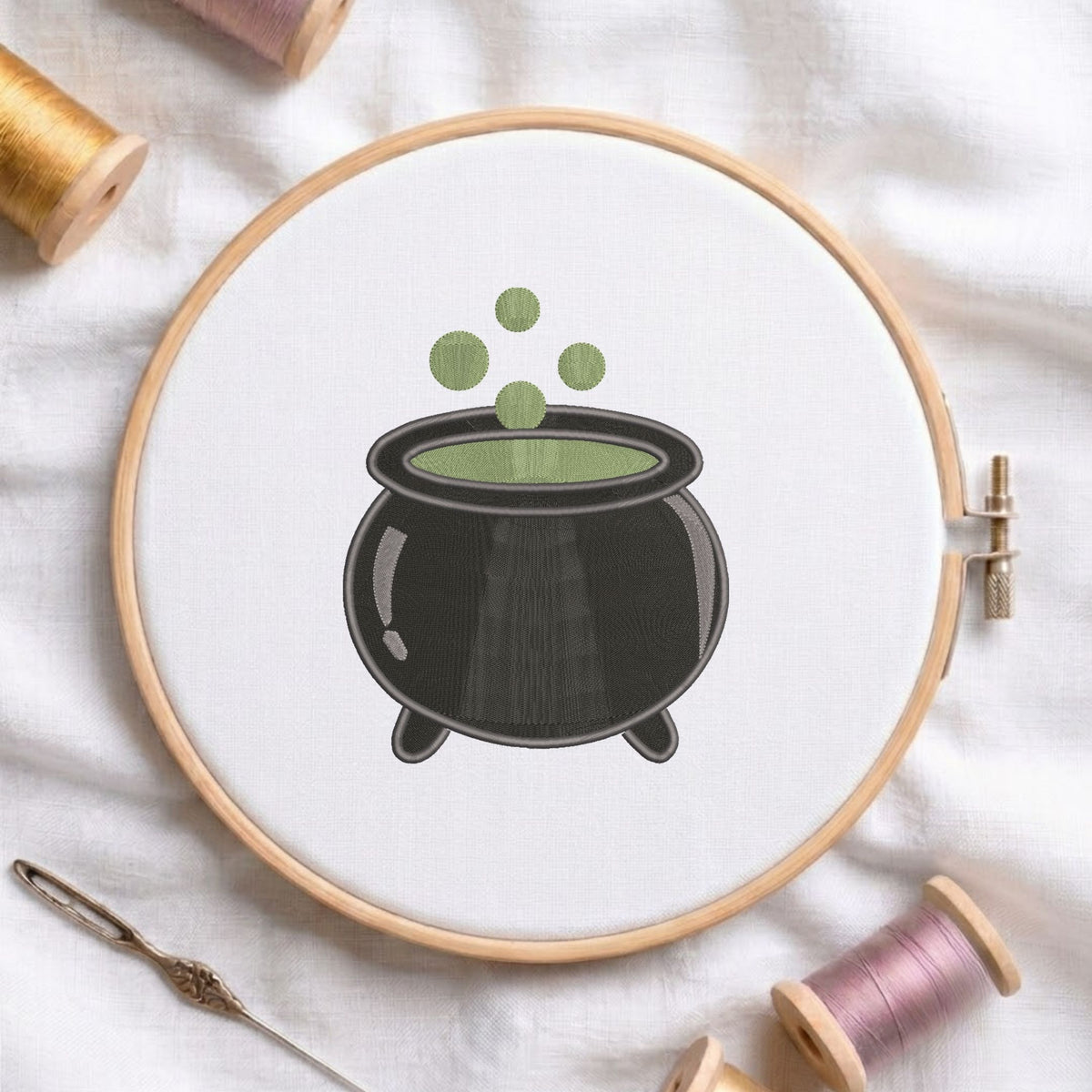Halloween witch cauldron machine embroidery design featuring a black bubbling cauldron with green potion, displayed inside a round wooden hoop on white linen fabric alongside thread spools and a needle.