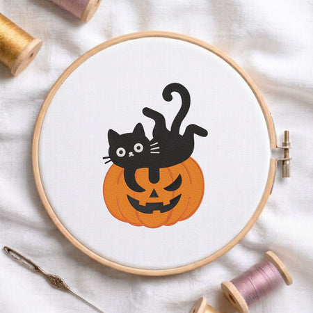 Cute black cat lying playfully on top of a carved jack-o-lantern pumpkin Halloween machine embroidery design shown in a wooden embroidery hoop on white fabric with thread spools and a needle