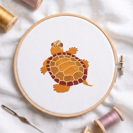  Turtle machine embroidery design displayed in a wooden hoop on white fabric with thread spools and a needle   