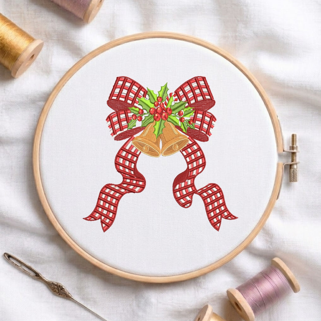 A circular embroidery hoop on white fabric, featuring a festive Christmas design with two gold bells, red gingham ribbon bows, and green holly with red berries. A needle and spools of thread are arranged around it on a white fabric background.
