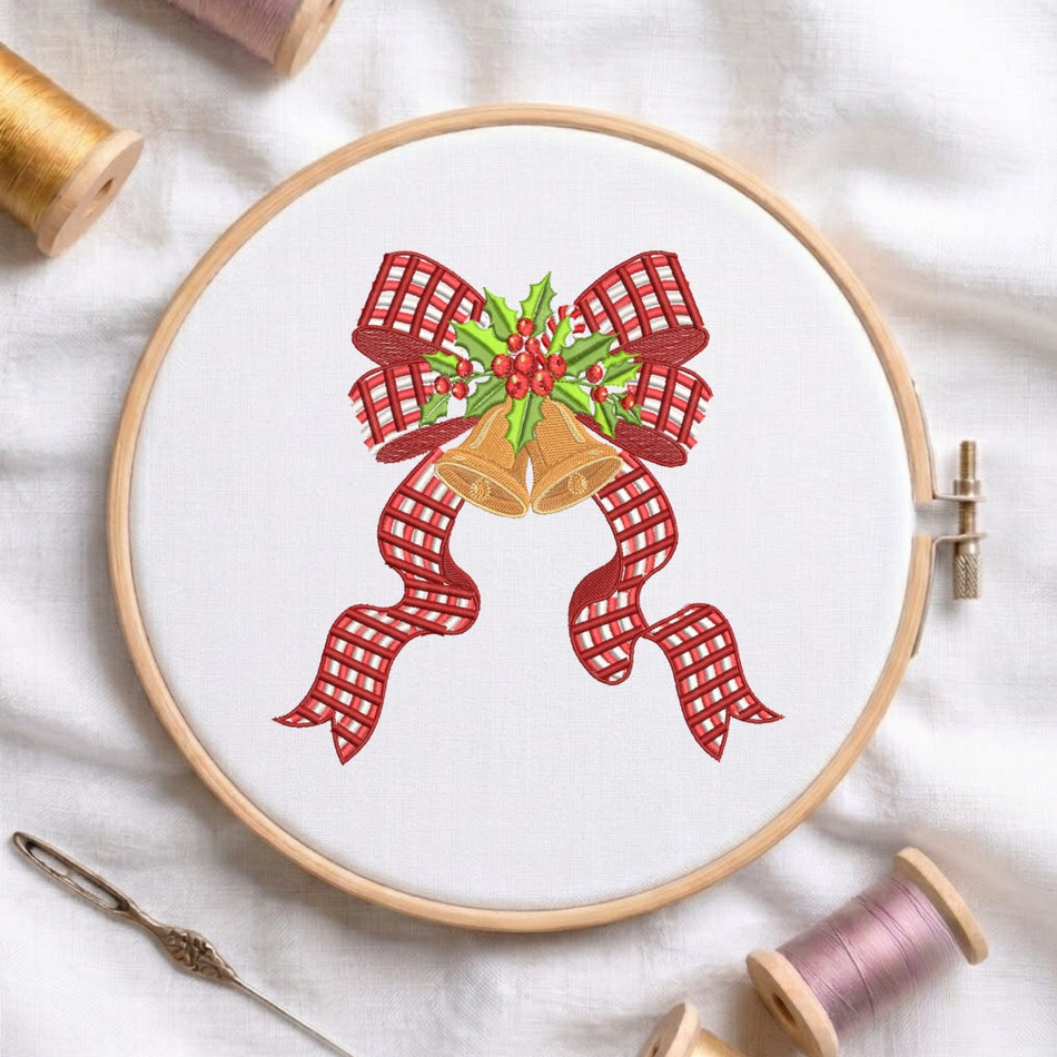 A circular embroidery hoop on white fabric, featuring a festive Christmas design with two gold bells, red gingham ribbon bows, and green holly with red berries. A needle and spools of thread are arranged around it on a white fabric background.