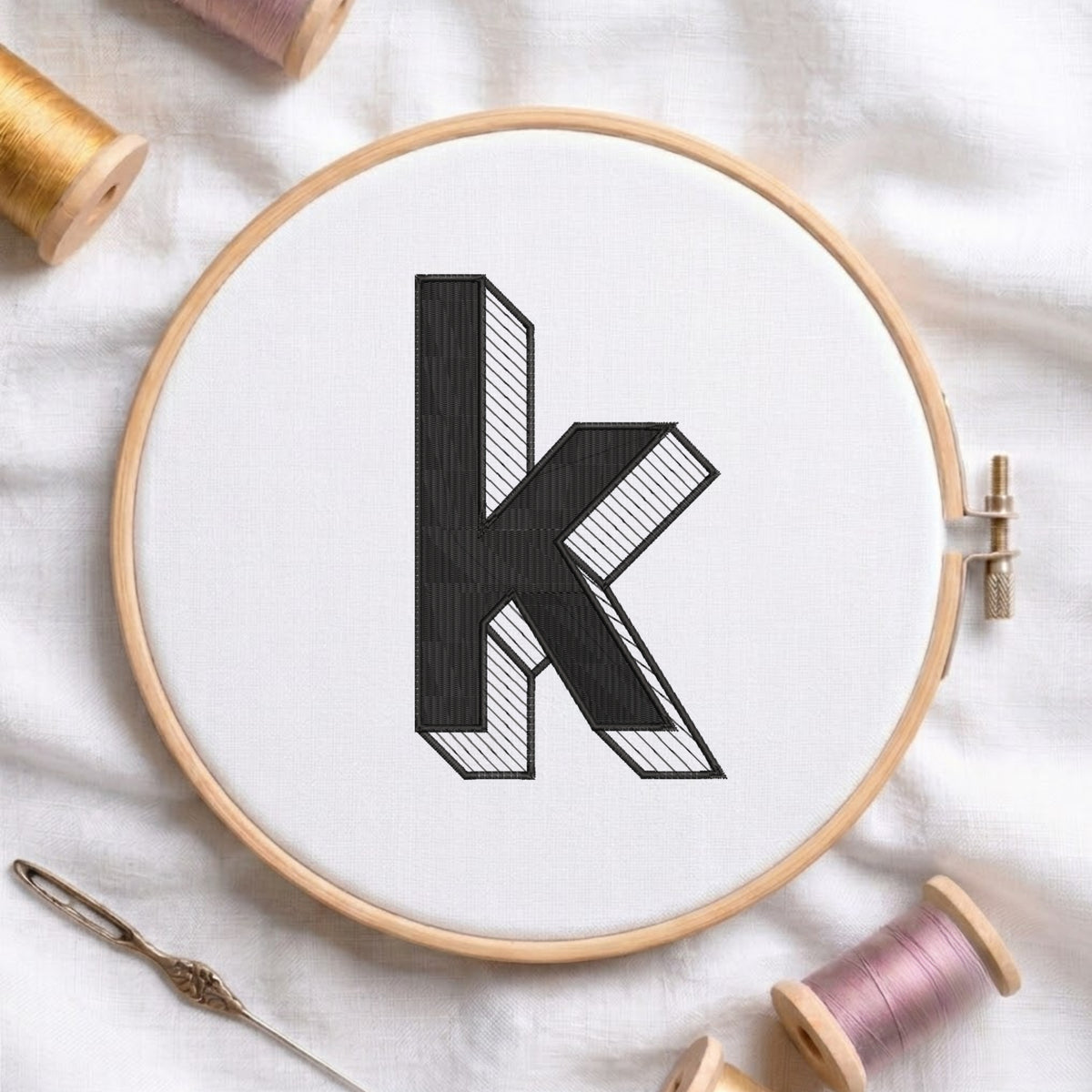 A wooden embroidery hoop holding white fabric with a large black letter 'k' stitched in a bold, stylized font. The hoop is photographed on a white fabric surface with spools of thread and a needle nearby.