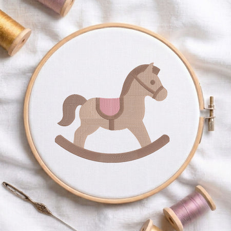 A circular embroidery hoop holding white fabric with a stylized rocking horse design in tan, brown, and pink. The hoop is photographed on a white fabric surface with spools of thread and a needle around it.
