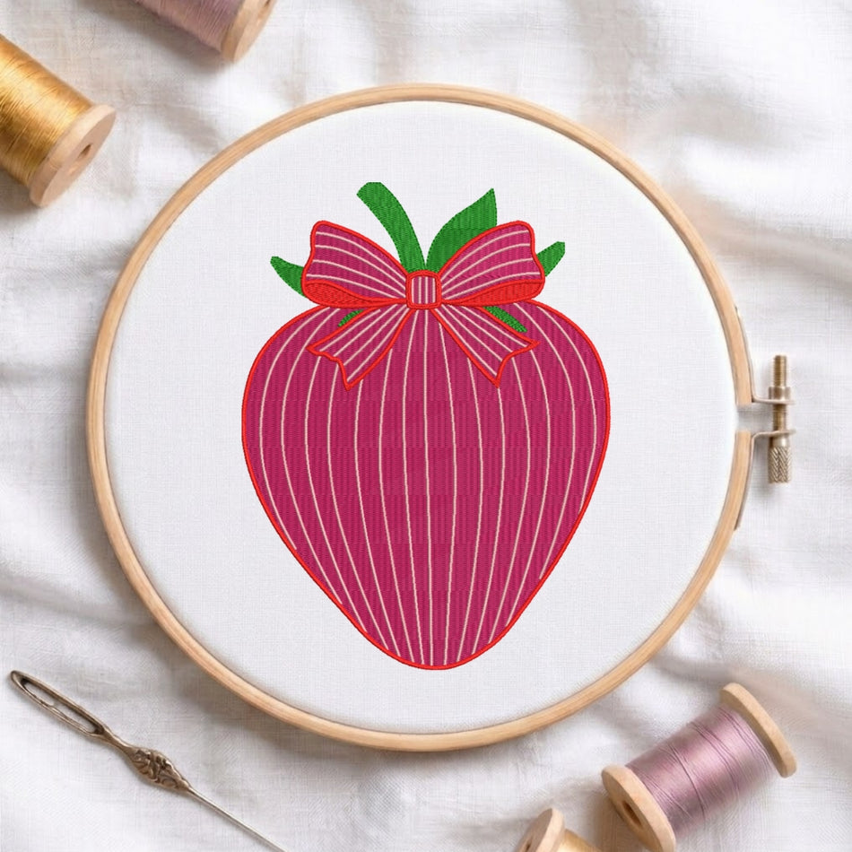 A wooden embroidery hoop holding white fabric with a stylized pink striped strawberry design topped with green leaves and a pink bow. Spools of thread, a needle, and soft fabric are arranged around it in a styled flat-lay.