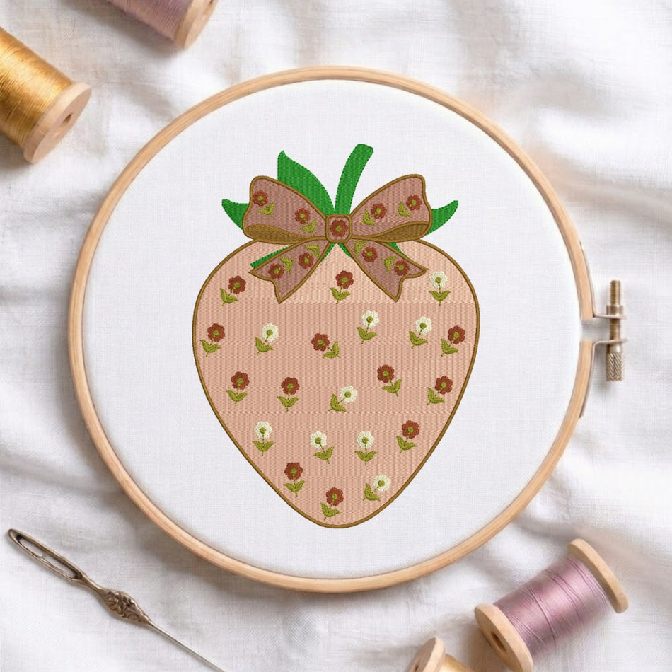 A wooden embroidery hoop holds white fabric with a stitched strawberry design. The strawberry has a peach-pink striped body, small brown and white flowers, green leaves, and a brown bow on top.