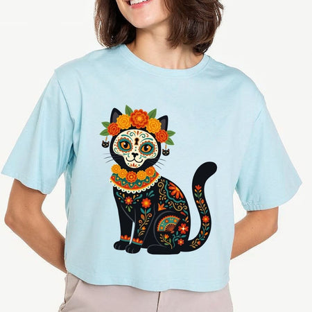 A woman wearing a light blue cropped t-shirt with a colorful Dia de los Muertos cat graphic, paired with beige pants, against a plain white background.