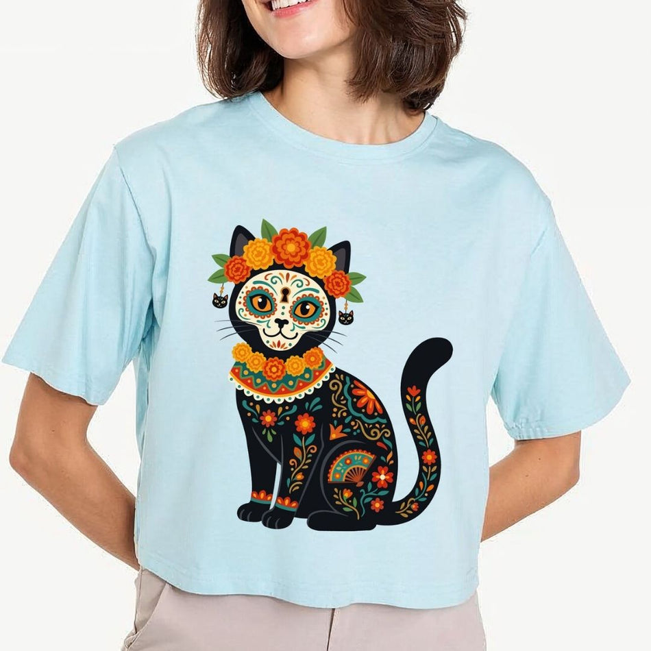 A woman wearing a light blue cropped t-shirt with a colorful Dia de los Muertos cat graphic, paired with beige pants, against a plain white background.