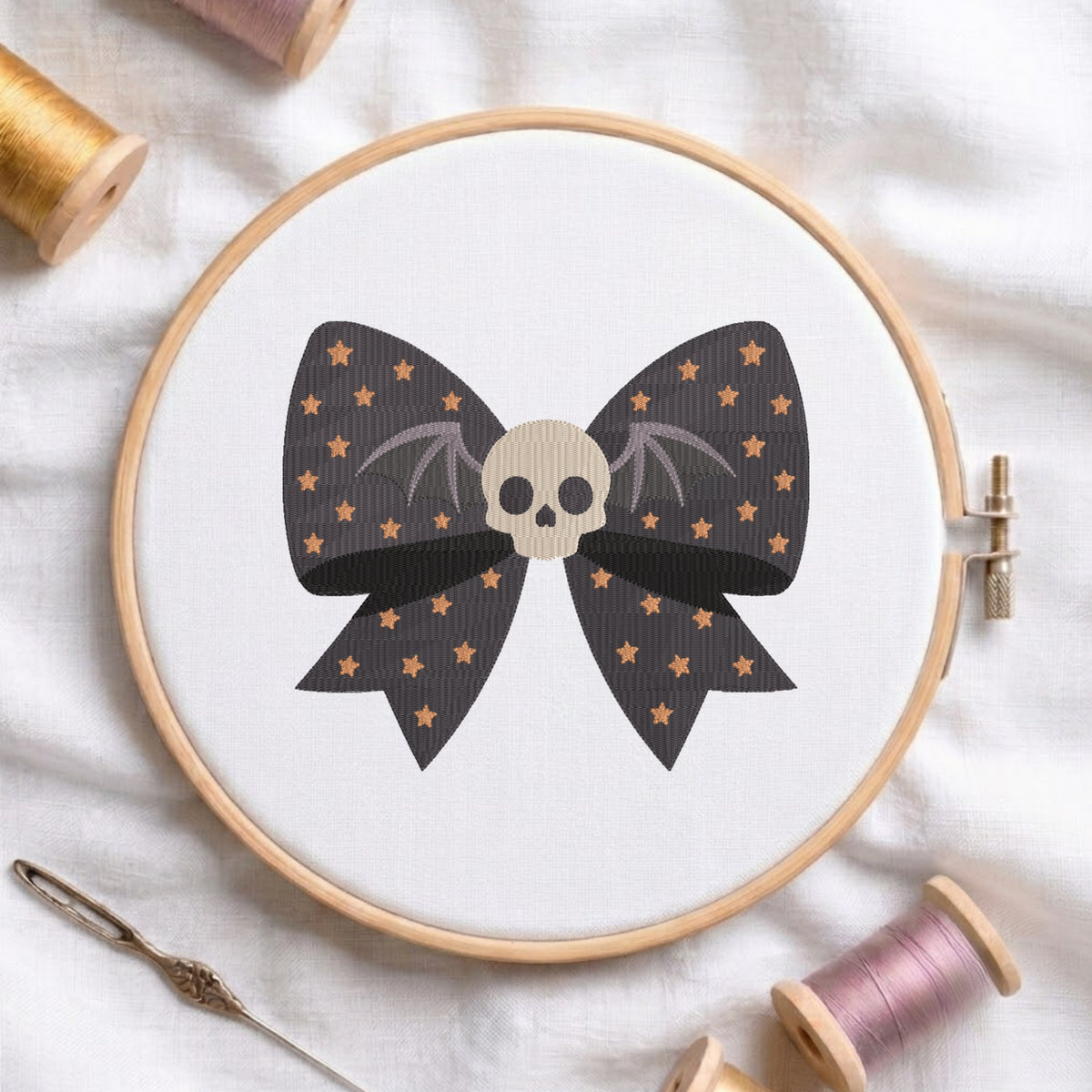 A white fabric stretched in a wooden embroidery hoop, featuring an embroidered dark bow patterned with gold stars, bat wings, and a centered beige skull.