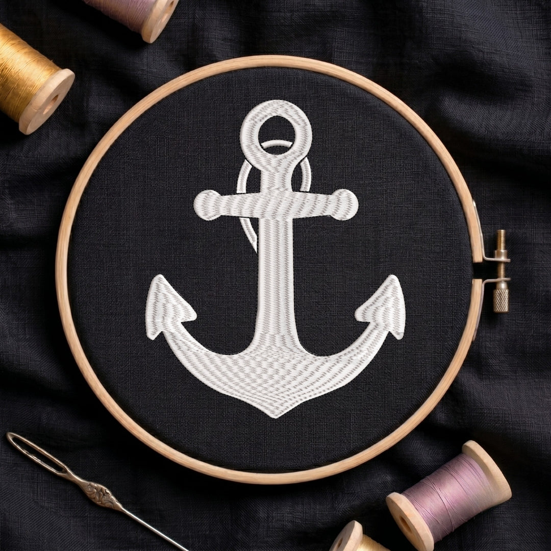 Embroidery hoop with a white anchor design on black fabric, surrounded by spools of thread.