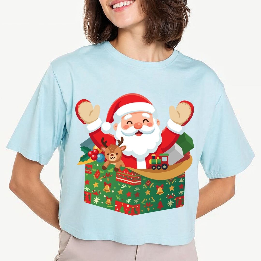 A woman wearing a light blue short-sleeve t-shirt with a colorful graphic of Santa Claus popping out of a Christmas gift box, set against a plain light gray background.