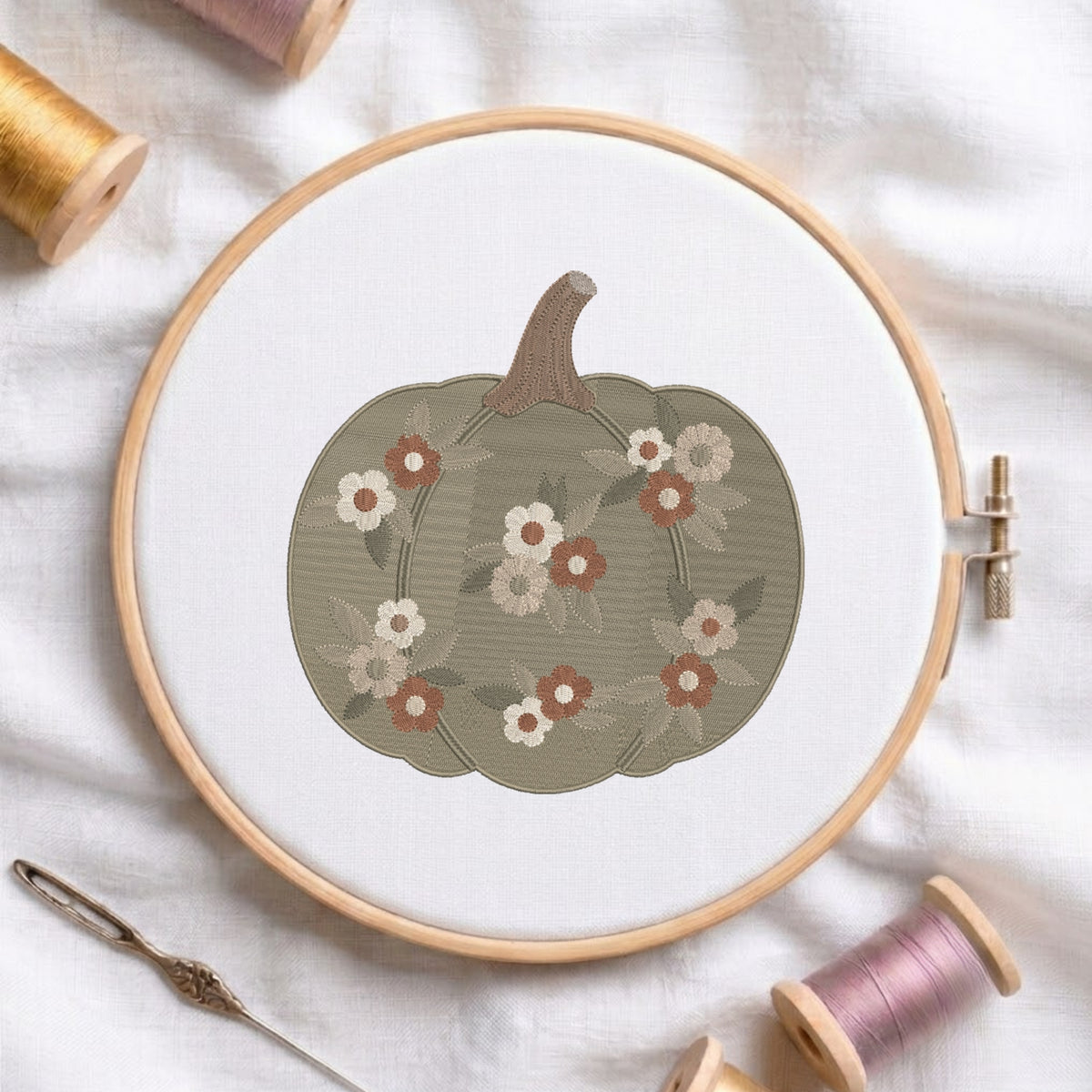 A circular embroidery hoop holds white fabric with a stitched pumpkin design in muted olive green, accented with cream, rust, and gray flowers. A metal needle lies beside the hoop, with spools of thread around it on a white fabric background.
