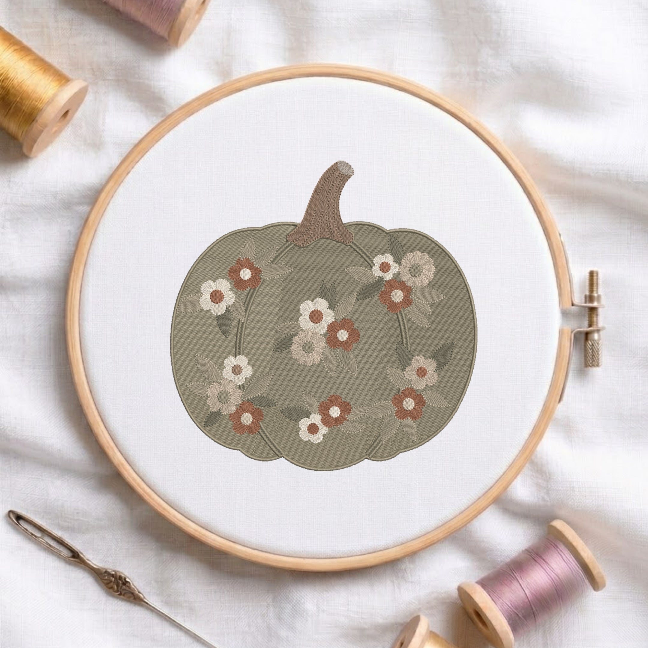 A circular embroidery hoop holds white fabric with a stitched pumpkin design in muted olive green, accented with cream, rust, and gray flowers. A metal needle lies beside the hoop, with spools of thread around it on a white fabric background.