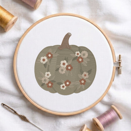 A circular embroidery hoop holds white fabric with a stitched pumpkin design in muted olive green, accented with cream, rust, and gray flowers. A metal needle lies beside the hoop, with spools of thread around it on a white fabric background.