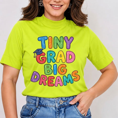 A woman wearing a bright lime green mock neck t-shirt with a colorful 'TINY GRAD BIG DREAMS' graphic print, paired with blue jeans.