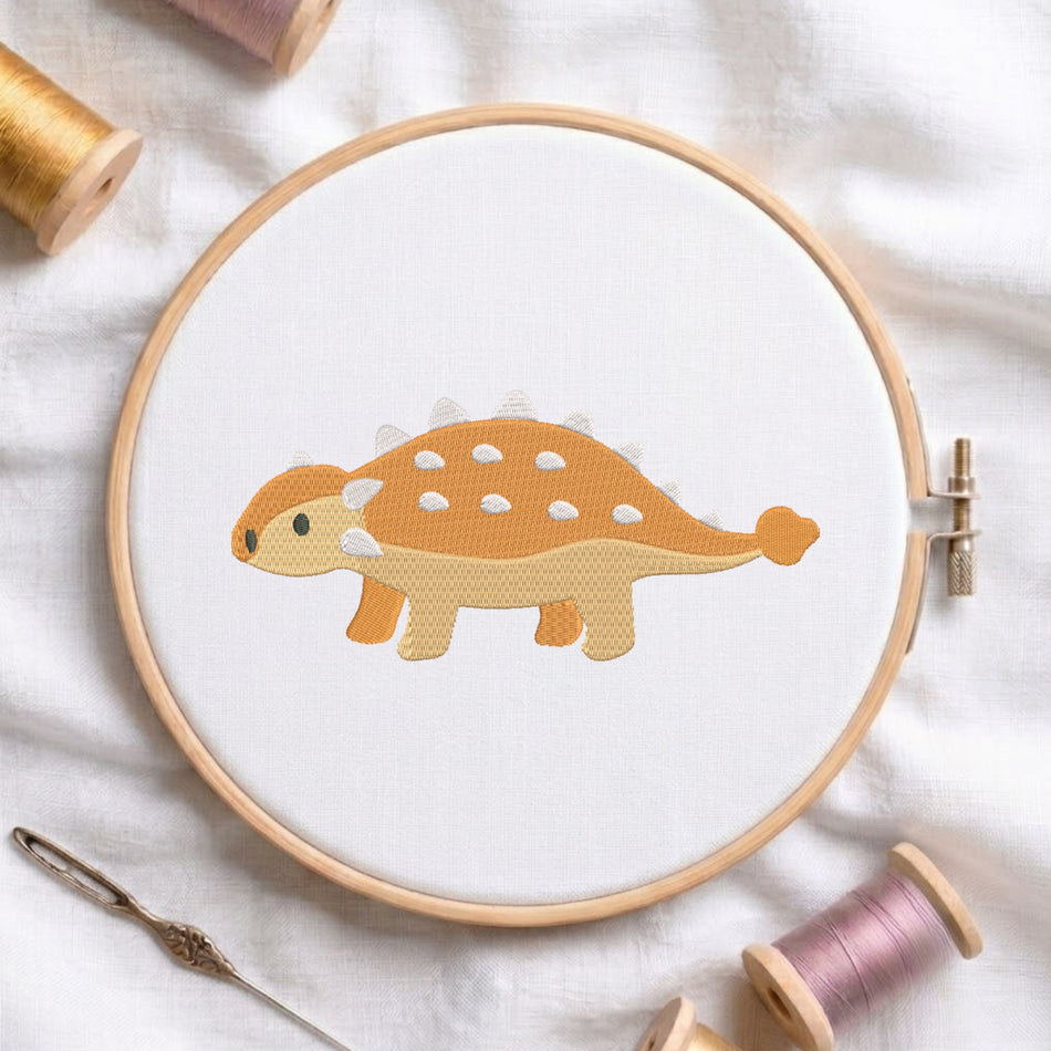 A wooden embroidery hoop holds a white fabric with a stitched cartoon dinosaur in orange, beige, and white. A needle and thread spools are arranged around it on a white fabric background.
