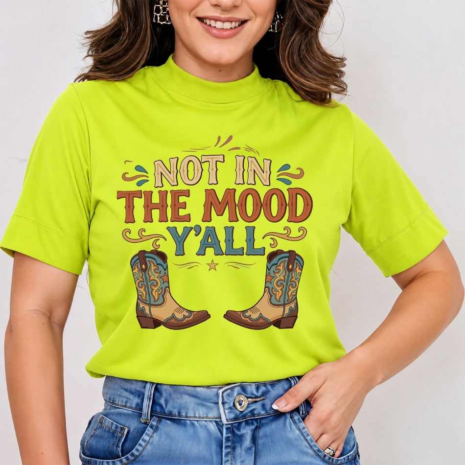 A woman wearing a bright lime green mock-neck t-shirt with a western-themed graphic that reads 'NOT IN THE MOOD Y'ALL' above a pair of cowboy boots, paired with blue jeans.