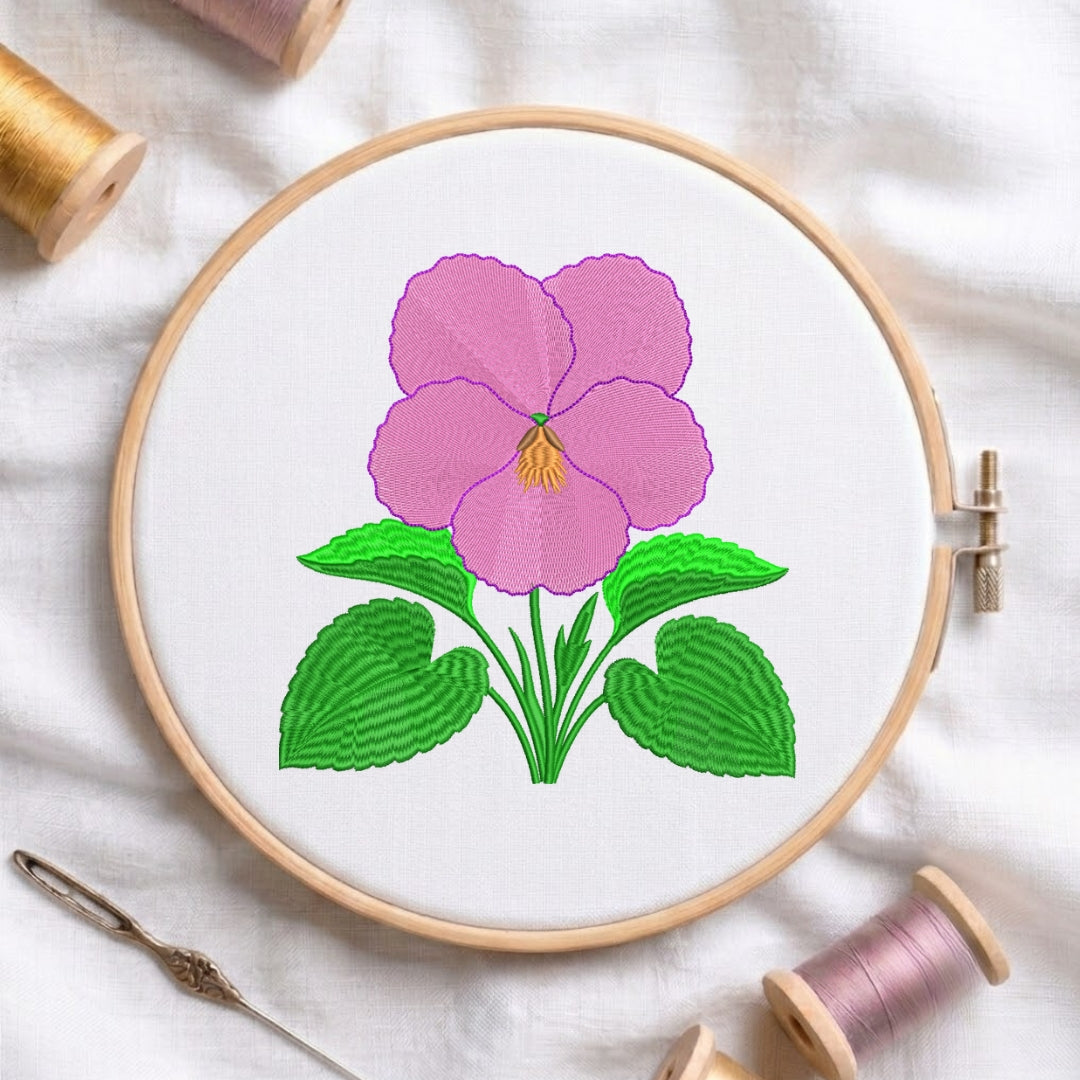 A pink pansy embroidery design centered in a wooden hoop, accompanied by a needle and two spools of thread on a white fabric background.