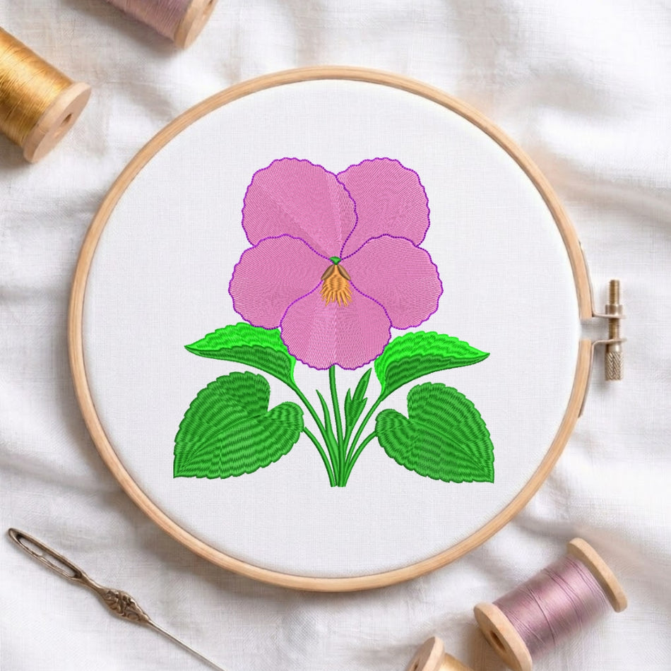 A pink pansy embroidery design centered in a wooden hoop, accompanied by a needle and two spools of thread on a white fabric background.