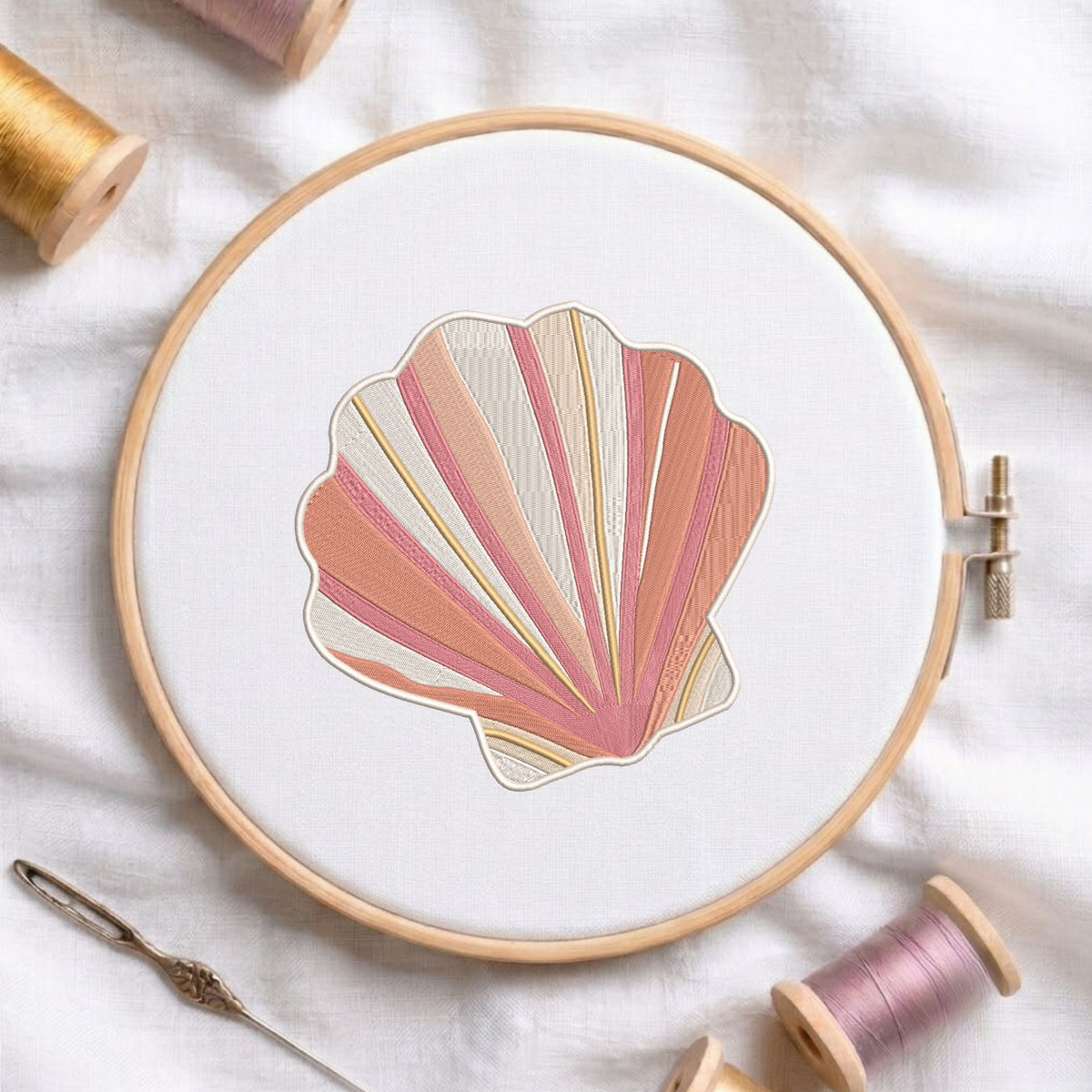 Pink and beige coastal seashell machine embroidery design displayed inside a round wooden hoop on white fabric alongside thread spools and a needle.