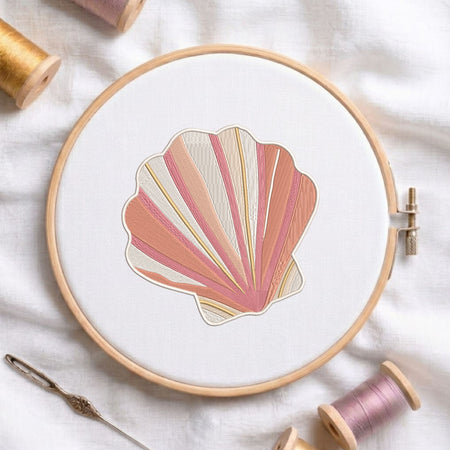 Pink and beige coastal seashell machine embroidery design displayed inside a round wooden hoop on white fabric alongside thread spools and a needle.