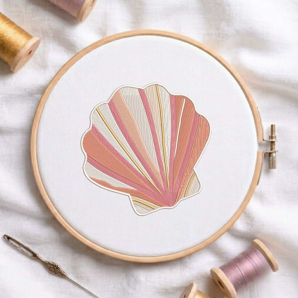 Pink and beige coastal seashell machine embroidery design displayed inside a round wooden hoop on white fabric alongside thread spools and a needle.