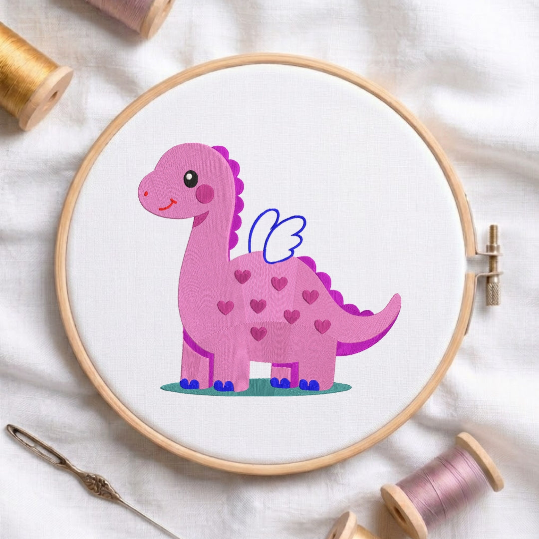 A circular embroidery hoop holding white fabric with a cute pink dinosaur design decorated with heart patterns, blue wing details, and a small green base. The hoop is styled on a white fabric background with spools of thread and a needle.