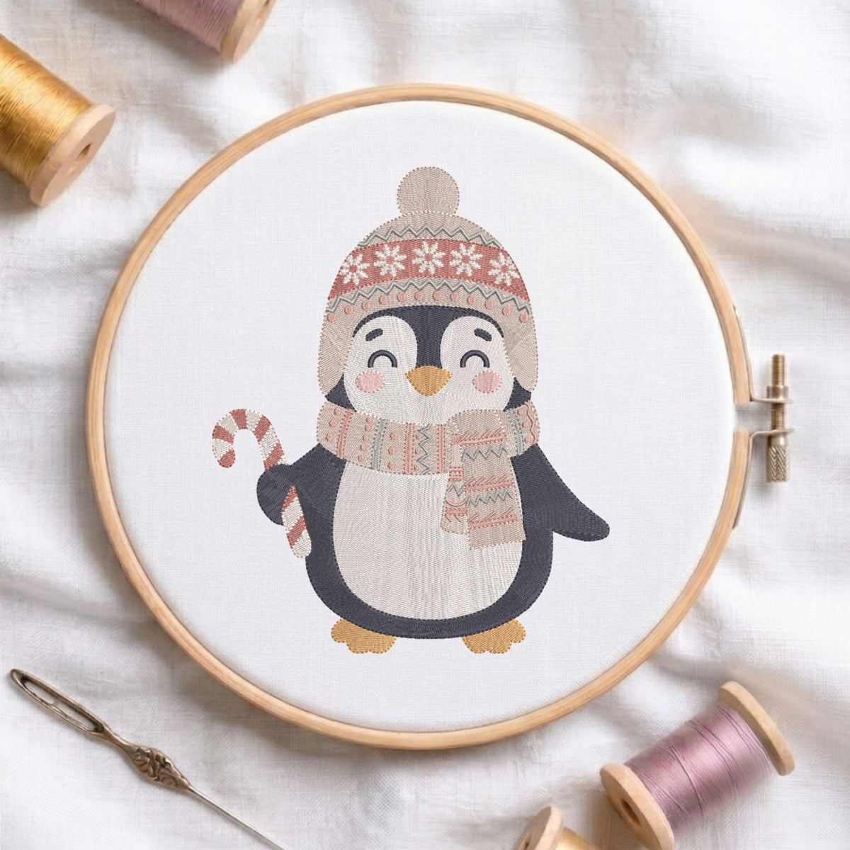 A circular embroidery hoop holds a white fabric with a stitched penguin wearing a knit hat and scarf in soft beige, pink, black, and orange tones. The hoop is styled on white fabric with spools of thread and a needle around it.