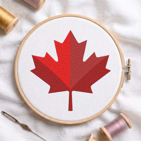 A circular embroidery hoop holding white fabric with a stylized red maple leaf design, surrounded by spools of thread and a needle on a white fabric background.