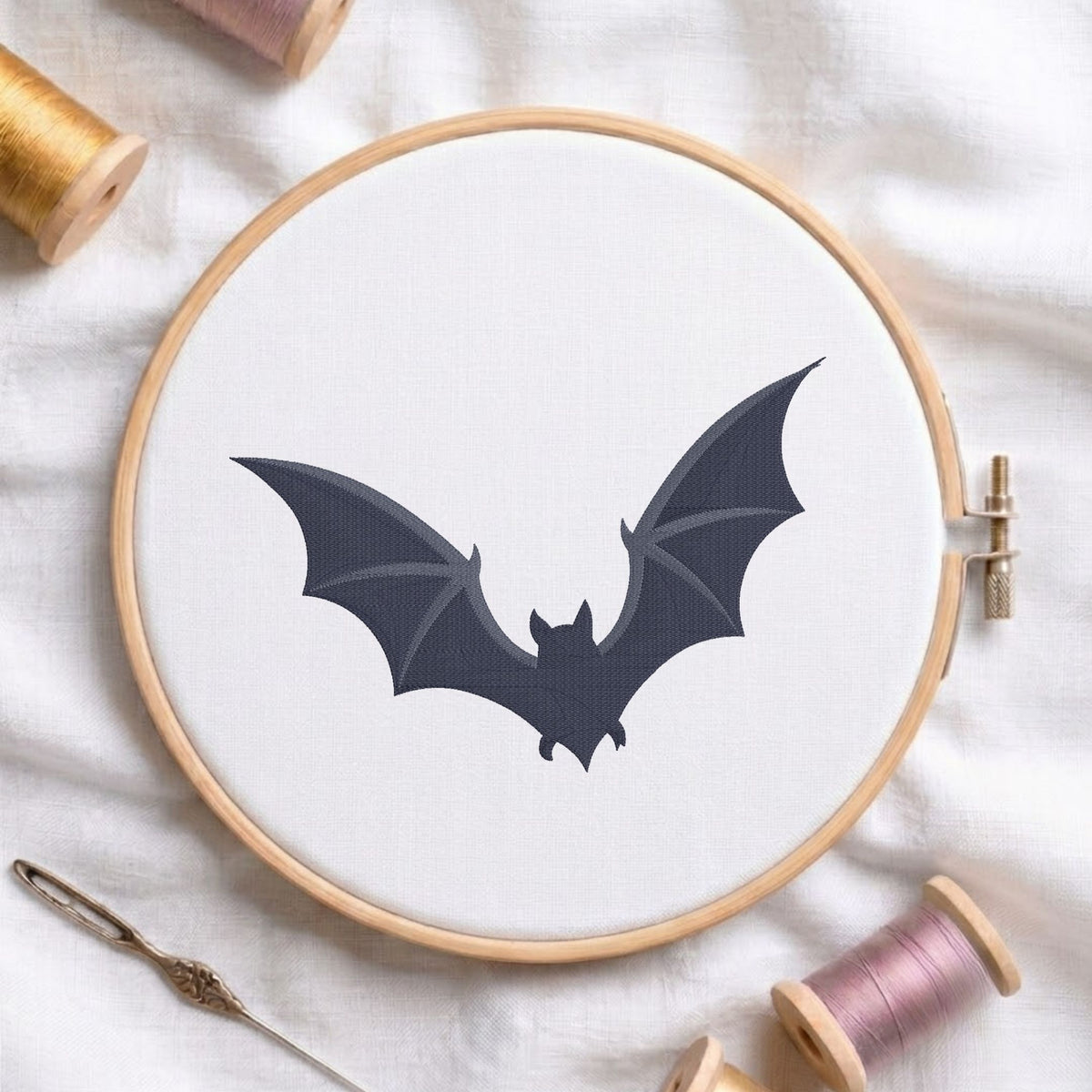 A bat silhouette design displayed inside a wooden embroidery hoop on white fabric, surrounded by spools of thread and a needle.