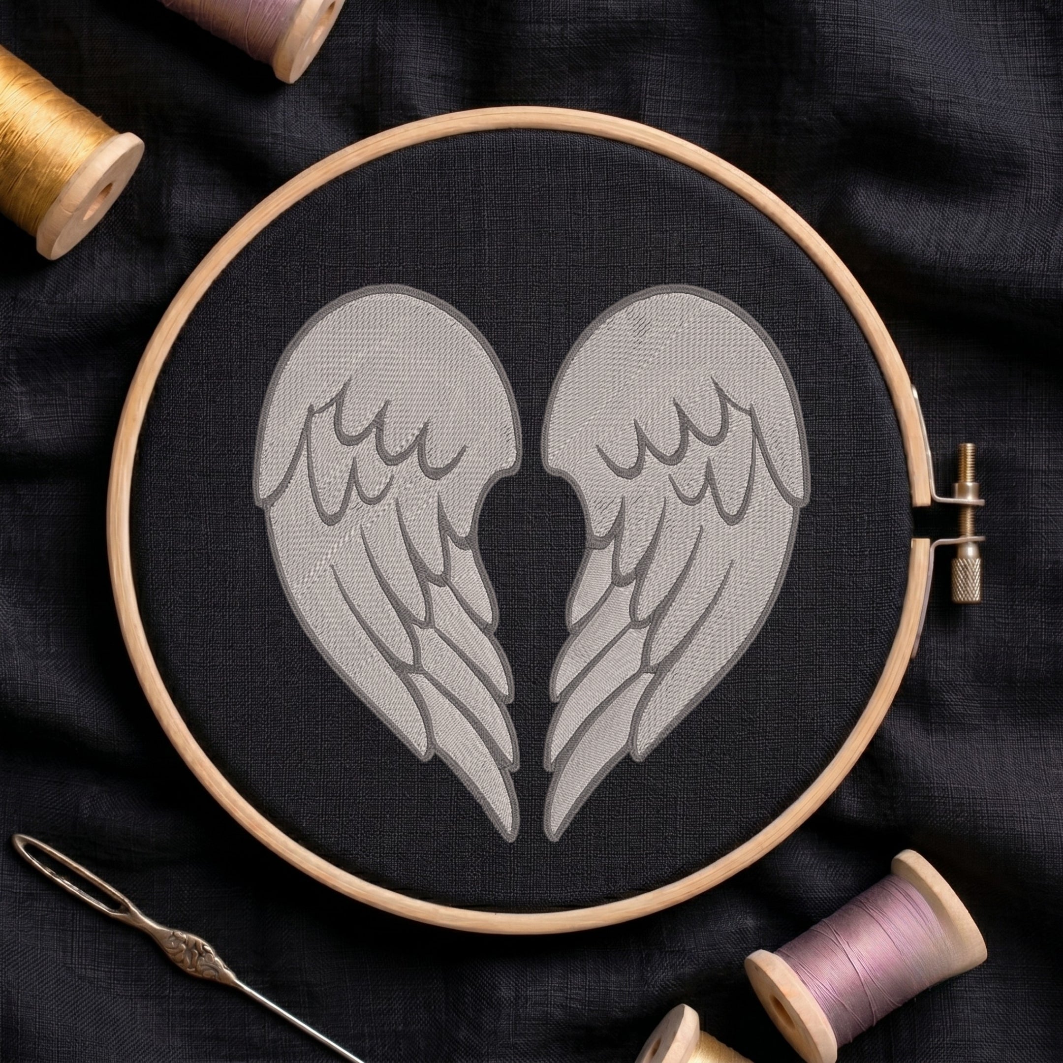 A circular embroidery hoop holding black fabric with a stitched design of two gray angel wings facing inward. The hoop is photographed on dark fabric with spools of thread and a needle around it.
