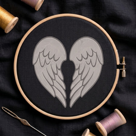 A circular embroidery hoop holding black fabric with a stitched design of two gray angel wings facing inward. The hoop is photographed on dark fabric with spools of thread and a needle around it.