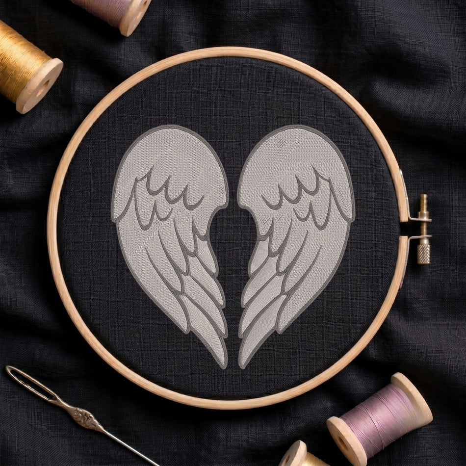 A circular embroidery hoop holding black fabric with a stitched design of two gray angel wings facing inward. The hoop is photographed on dark fabric with spools of thread and a needle around it.