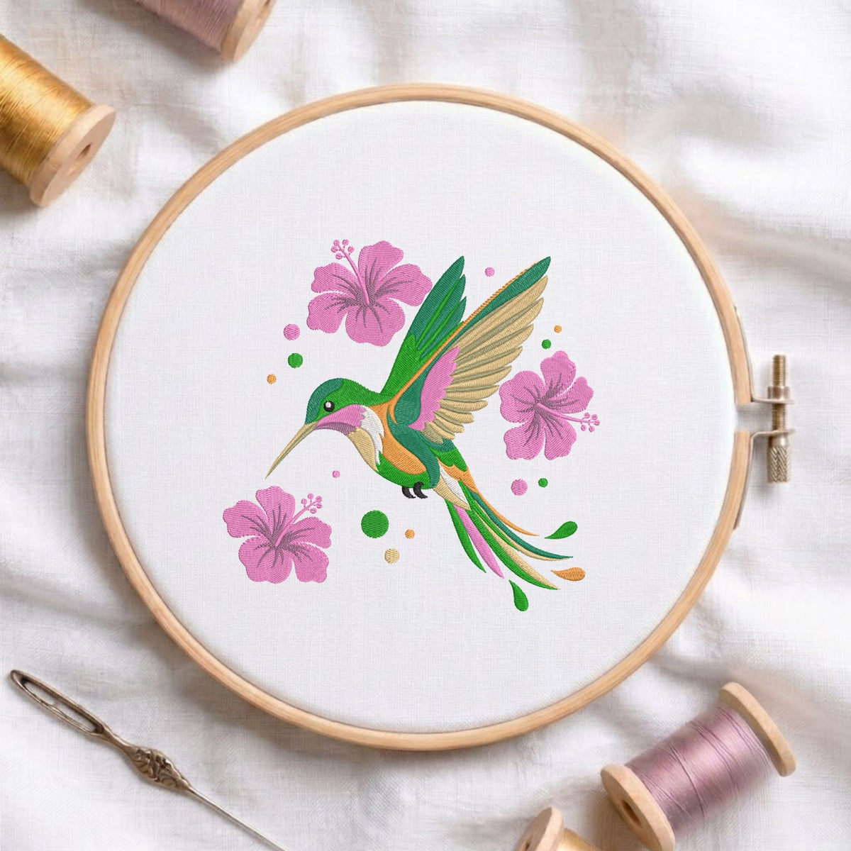 A round embroidery hoop holding white fabric with a colorful hummingbird and pink flowers design, surrounded by spools of thread and a needle on a white fabric background.