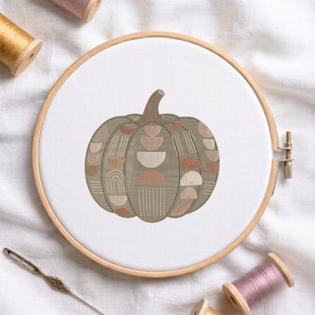 Geometric patterned pumpkin machine embroidery design displayed in a wooden hoop on white fabric with thread spools and a needle.   