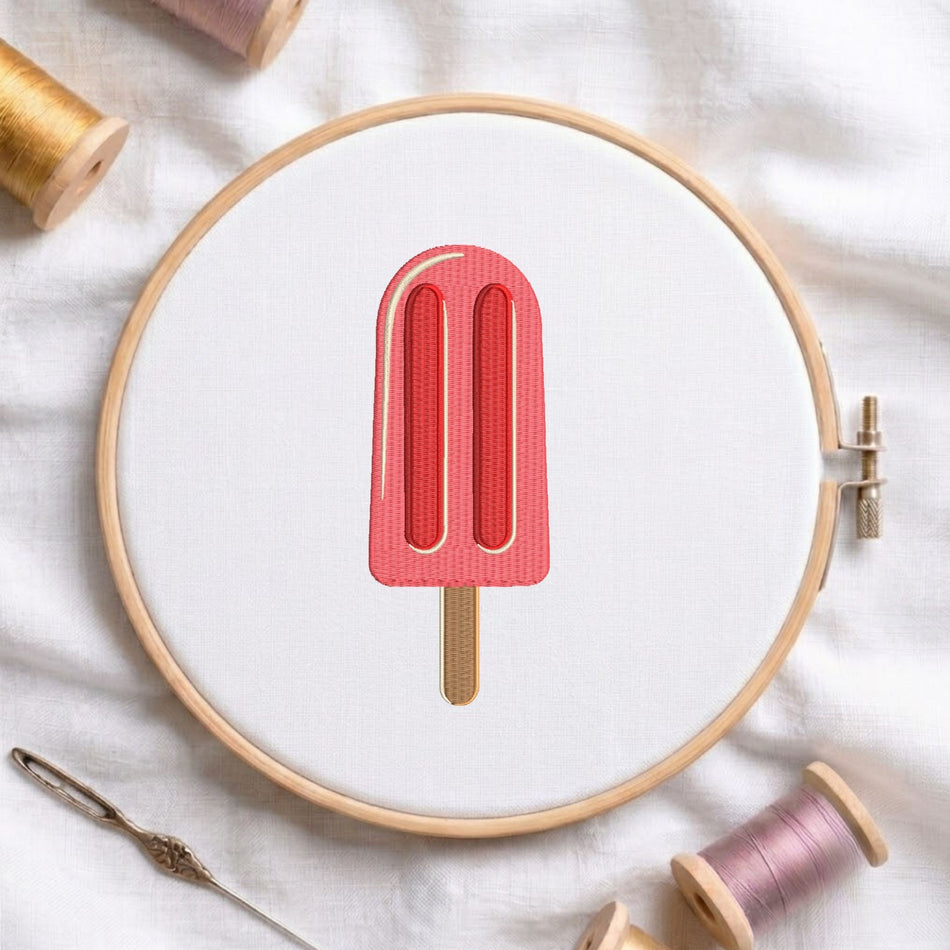 Pink ice cream popsicle with wooden stick machine embroidery design, displayed inside a round wooden hoop on white fabric with thread spools and a needle.   